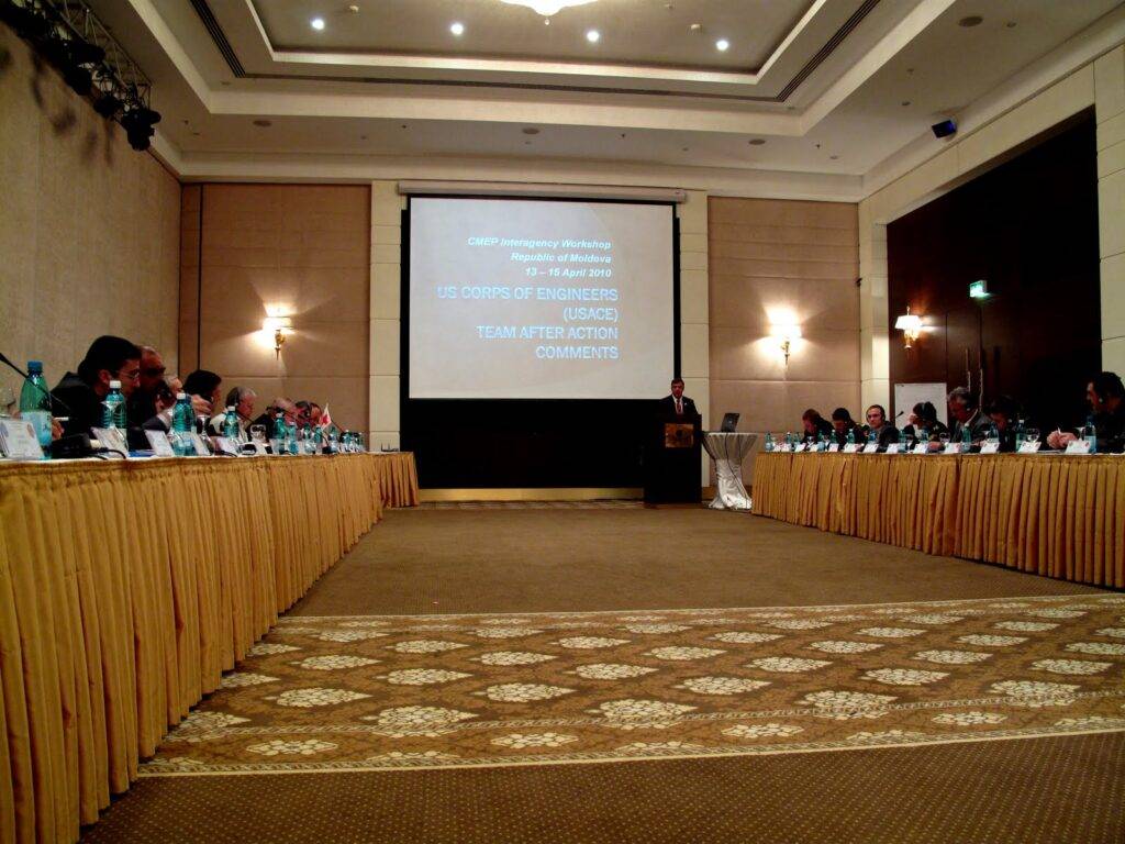 A large conference room with people seated around two long tables facing a screen. A presenter stands at a podium. The screen displays a workshop title and the text 
