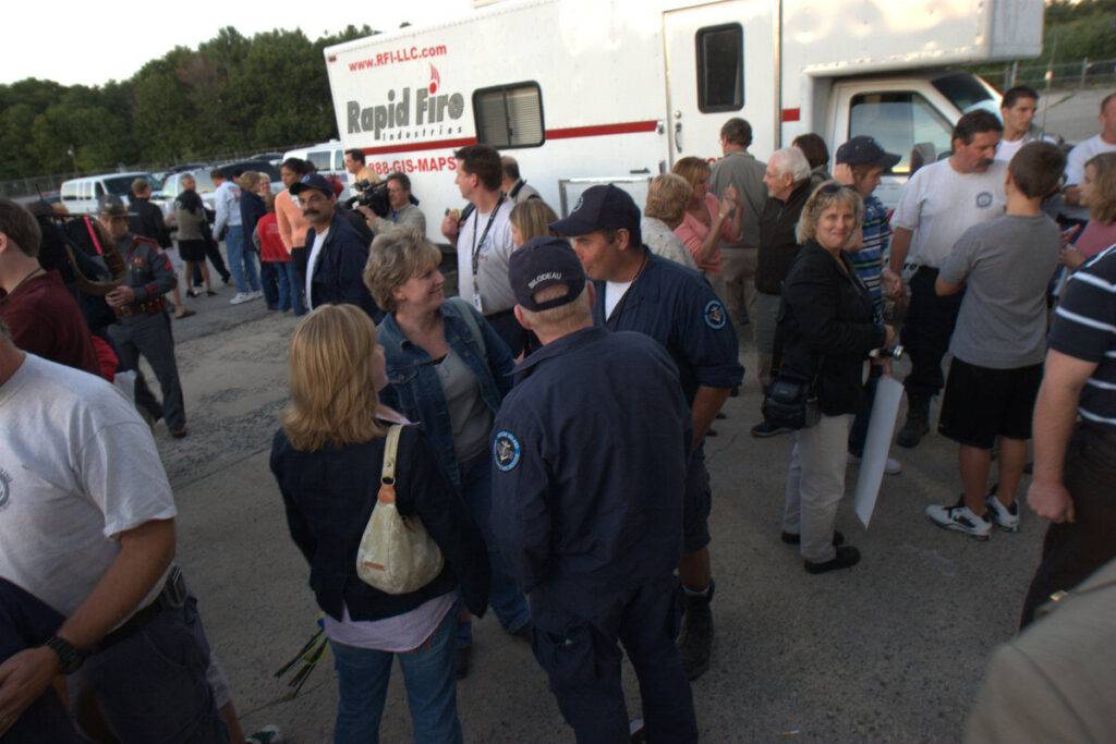 A group of people, including emergency workers in uniform, gather and talk outside near a white 