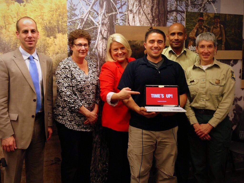 Six adults stand indoors, smiling at the camera. The man in the center holds a laptop displaying 