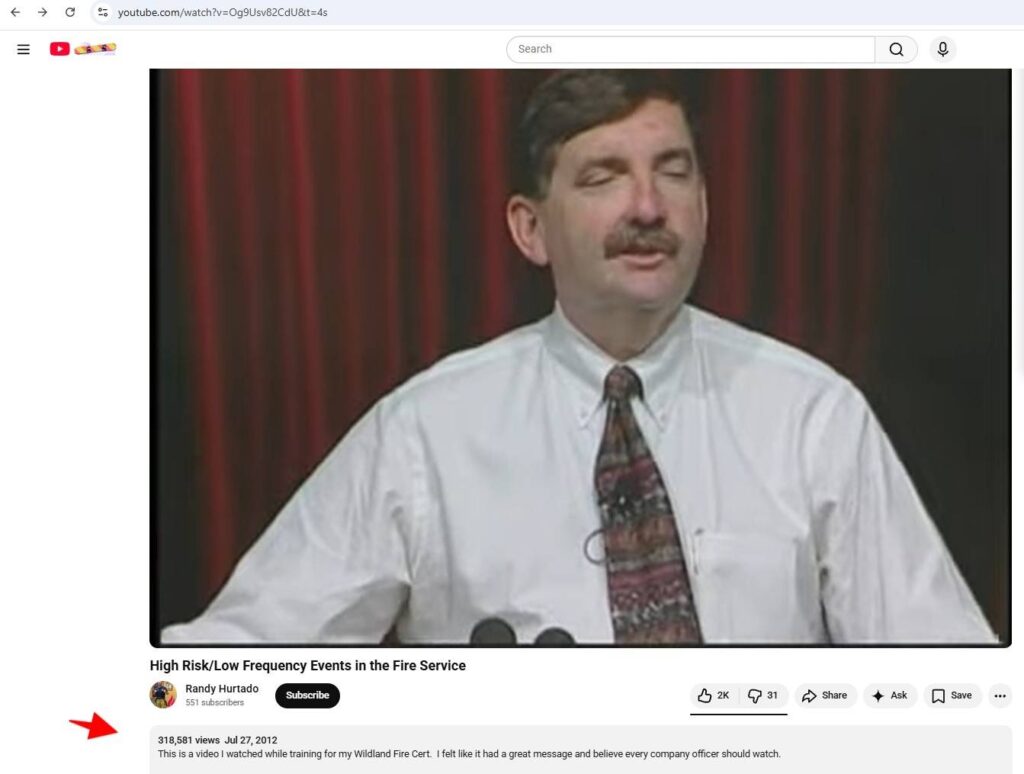 A man with a mustache in a white shirt and patterned tie stands giving a speech, with red and black vertical curtains in the background. The YouTube video is titled 