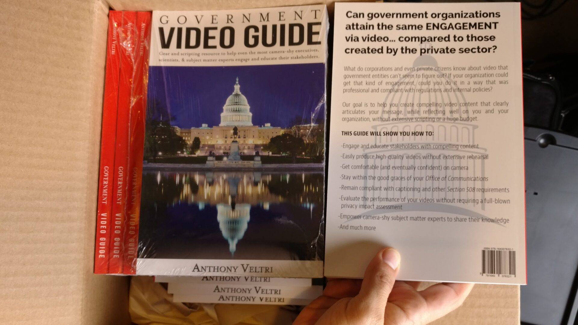 A hand holds a copy of the "Government Video Guide" by Anthony Veltri above a box containing more copies. The front cover shows the U.S. Capitol at night, while the back cover has text about engagement via video in government organizations.