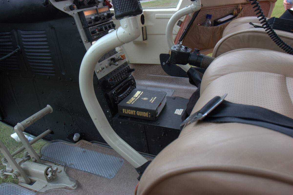 A close-up view of a helicopter cockpit interior, showing tan leather seats, flight controls, instrument panels, and a "Flight Guide" manual resting on a black box between the seats.
