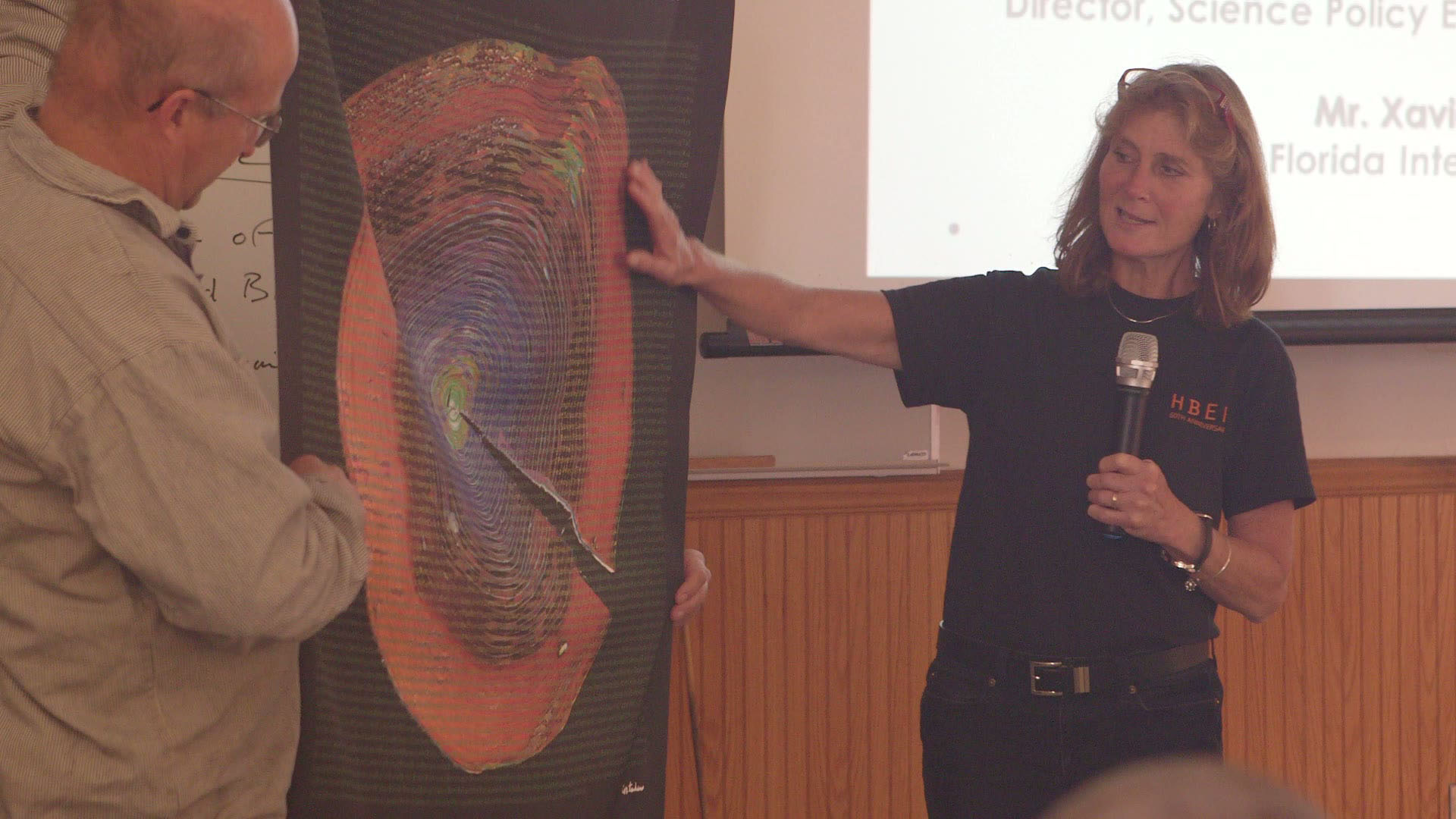 A woman holding a microphone stands beside a man and gestures to a large, colorful spiral diagram on a poster. The diagram features swirling red, orange, and green shapes with a pointed object in the center. They appear to be giving a presentation.
