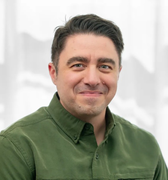 A man with short, dark hair and light skin smiles gently at the camera. He is wearing a moss-green button-up shirt. The background is softly blurred and light, creating a calm, neutral setting.