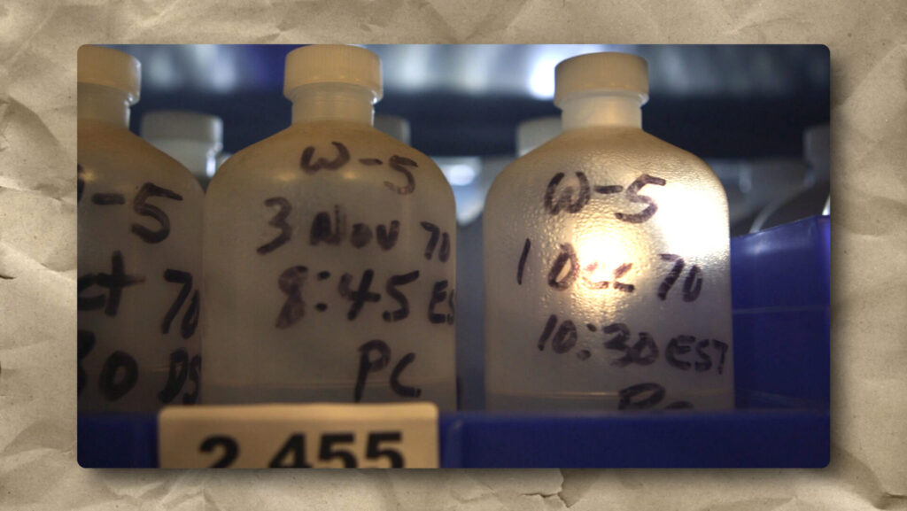 A close-up of frosty plastic bottles labeled by hand with dates and times, placed in a blue storage rack. The label “W-5” and dates like “3 Nov 70” and “10-2-70” are visible. The background is blurred, emphasizing the bottles.
