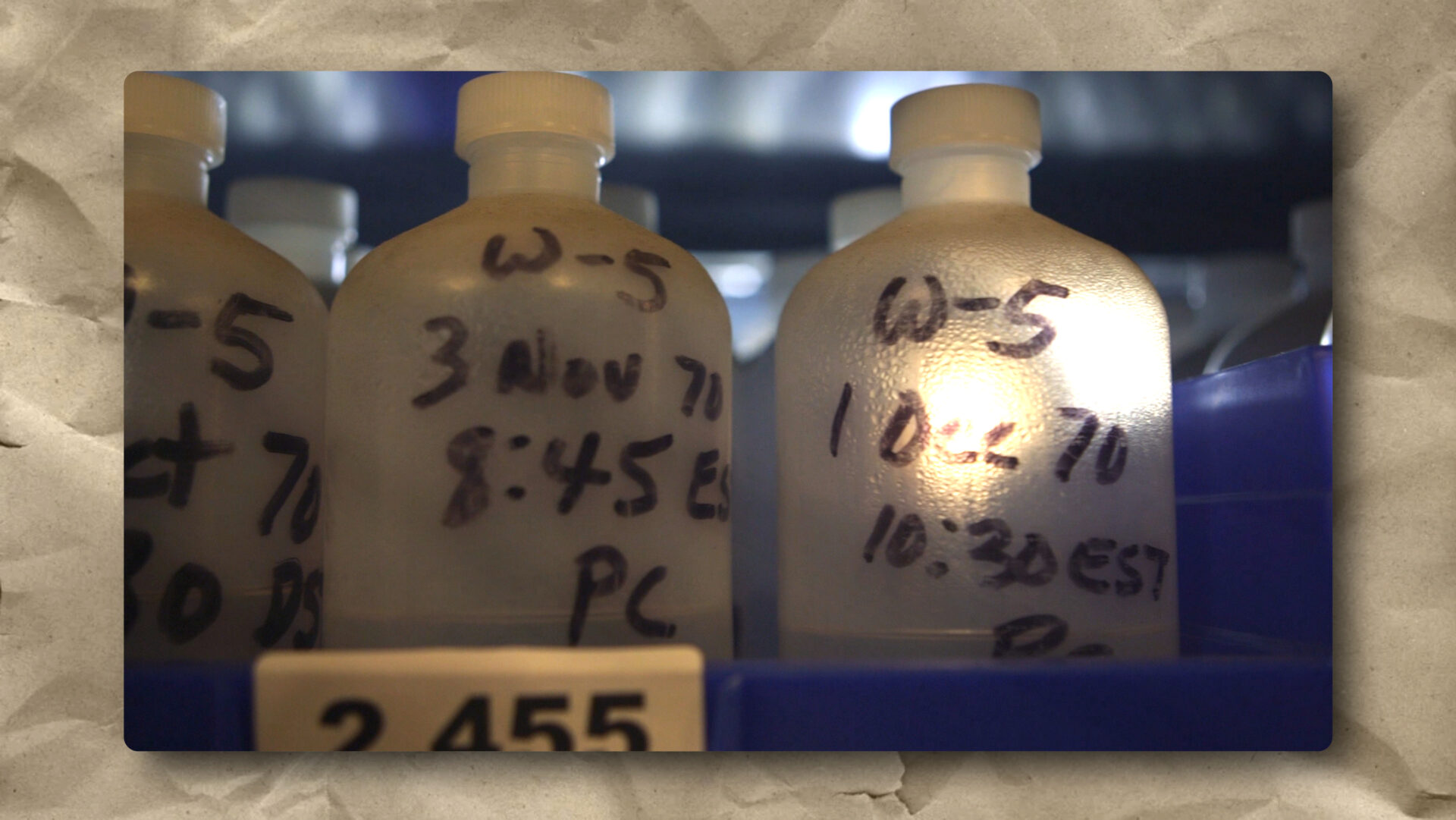 A close-up of frosty plastic bottles labeled by hand with dates and times, placed in a blue storage rack. The label “W-5” and dates like “3 Nov 70” and “10-2-70” are visible. The background is blurred, emphasizing the bottles.