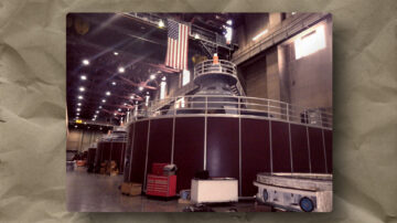 Interior of a large industrial facility with several massive, cylindrical machines lined up. An American flag hangs above them. The area is lit by rows of ceiling lights and scattered with equipment, boxes, and railings, giving a sense of scale and function.