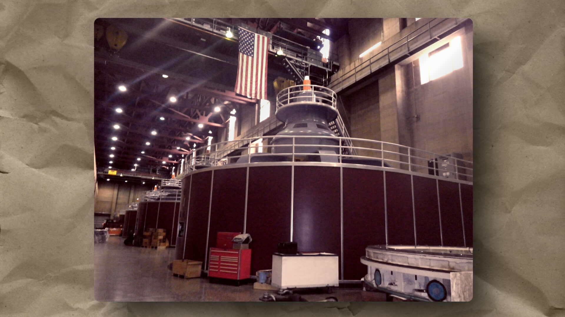 Interior of a large industrial facility with several massive, cylindrical machines lined up. An American flag hangs above them. The area is lit by rows of ceiling lights and scattered with equipment, boxes, and railings, giving a sense of scale and function.