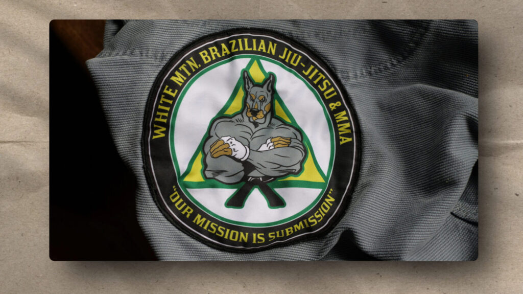 A fabric patch featuring a muscular, anthropomorphic dog in a gi with crossed arms, inside a green triangle. Yellow text reads “WHITE MTN. BRAZILIAN JIU-JITSU & MMA” and “OUR MISSION IS SUBMISSION” on a black background.