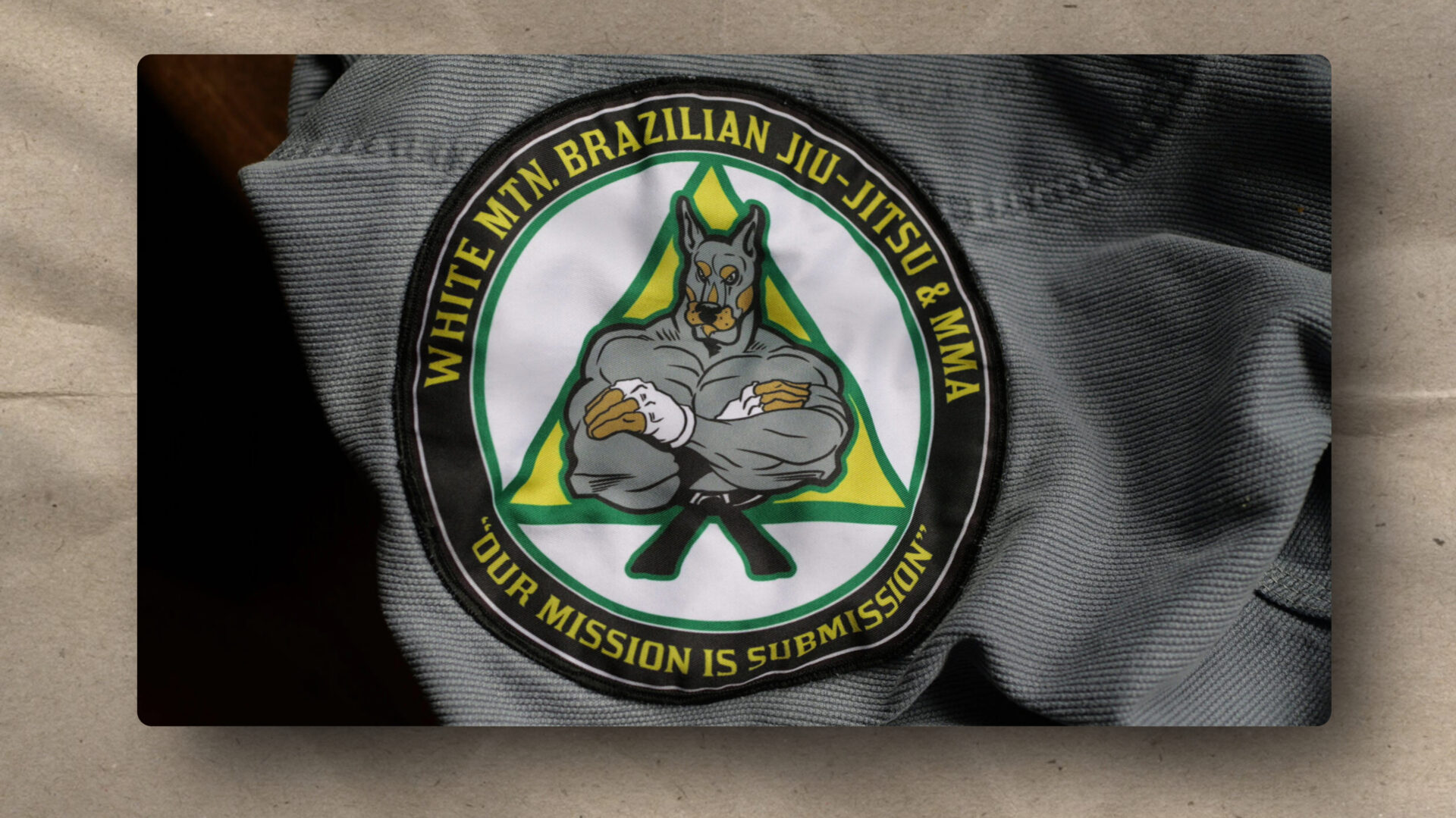 A fabric patch featuring a muscular, anthropomorphic dog in a gi with crossed arms, inside a green triangle. Yellow text reads “WHITE MTN. BRAZILIAN JIU-JITSU & MMA” and “OUR MISSION IS SUBMISSION” on a black background.