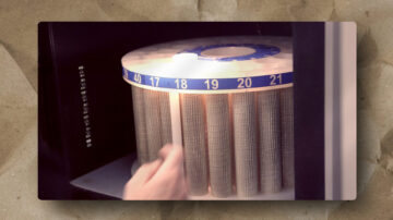 A close-up of a hand inserting or removing a rectangular card from a vintage cylindrical storage device with numbered slots from 17 to 21, each holding a metal mesh tube. The background shows a textured brown surface.