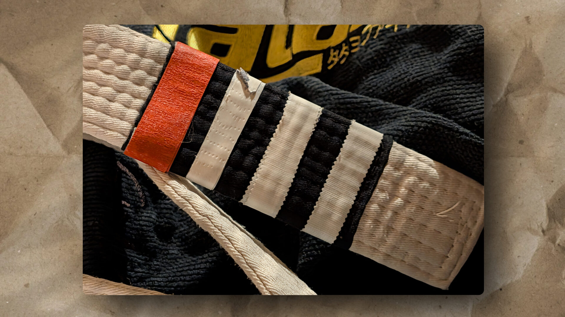 Close-up of a martial arts white belt with a red stripe and four black stripes, resting on a black uniform with gold text and Japanese characters. The textured fabric of the belt and uniform is visible against a crumpled brown paper background.