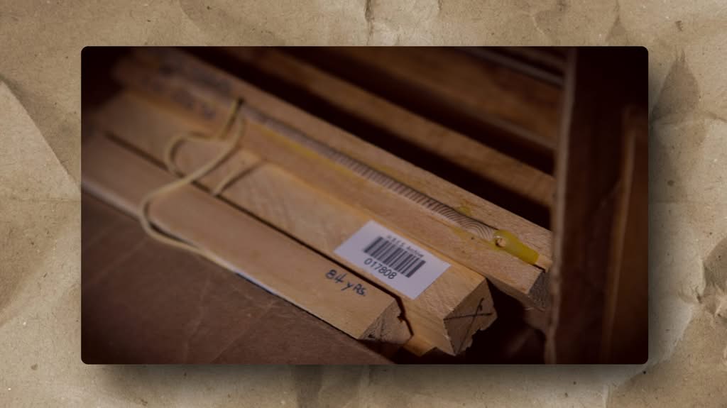 Close-up of wooden sticks with a barcode sticker, ruler markings, and string attached. The wood pieces are stacked on a cardboard surface, with handwritten numbers “8446” visible, and part of a yellow object beside them. Background is blurred.