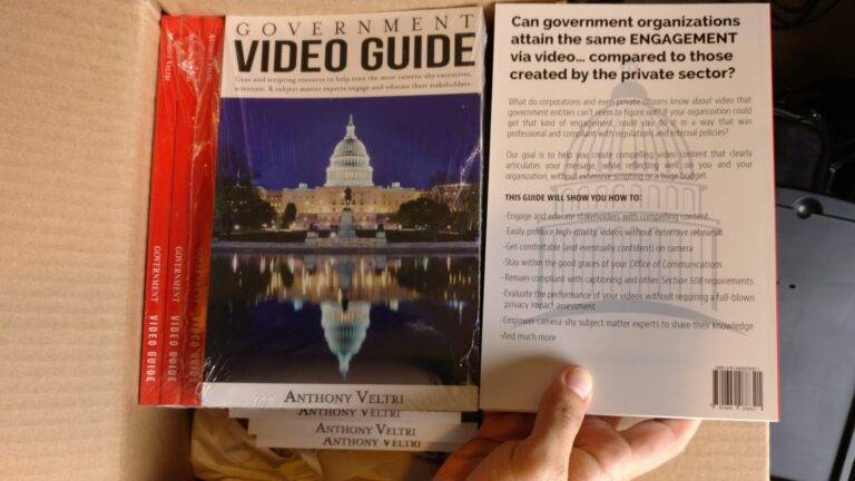 A hand holds a copy of the "Government Video Guide" by Anthony Veltri above a box containing more copies. The front cover shows the U.S. Capitol at night, while the back cover has text about engagement via video in government organizations.