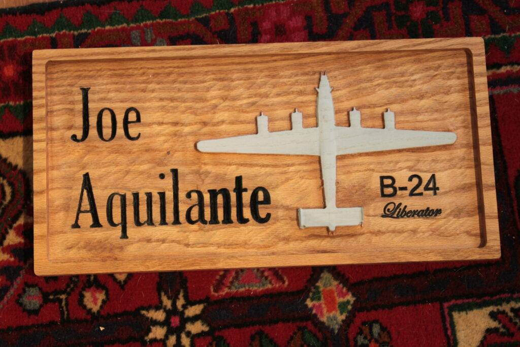 A wooden plaque with โJoe Aquilanteโ on the left, and a silhouette of a B-24 Liberator bomber on the right above the text โB-24 Liberator.โ The background is a patterned red and green carpet.