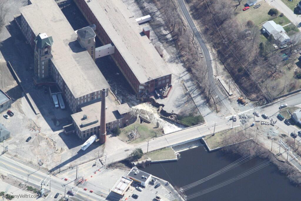 Aerial view of a large industrial building with a tall brick smokestack, next to a river. Part of the building and nearby road have collapsed, revealing debris and a large sinkhole. Construction vehicles and barriers surround the damaged area. Trees and houses are nearby.